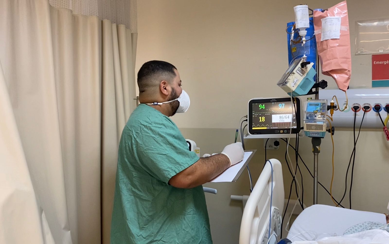 O enfermeiro está de costas vestindo avental verde e usando máscara de proteção cobrindo nariz e boca. Ele está com uma prancheta na mão anotando o que está no visor de uma máquina que mostra os batimentos cardíacos do paciente.